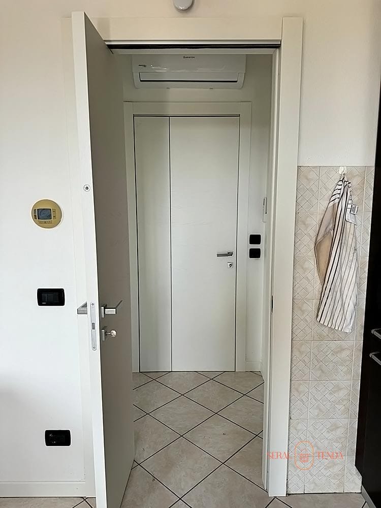 Porta Bertolotto Double bianca in un ingresso di un'abitazione a Secugnago. La porta aperta mostra un disimpegno con climatizzatore e una seconda porta interna bianca. Sistema completo di porte Bertolotto che valorizza gli spazi con design contemporaneo e finiture di qualità.