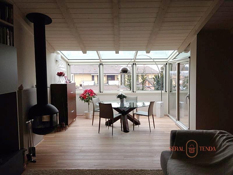 Veranda in alluminio con soffitto in legno bianco e ampie vetrate installata a Muzza di Cornegliano. Spazio luminoso arredato con tavolo rotondo in vetro, sedie bianche e stufa a legna. La veranda offre vista sul quartiere residenziale e trasforma il terrazzo in un elegante ambiente abitabile in ogni stagione.