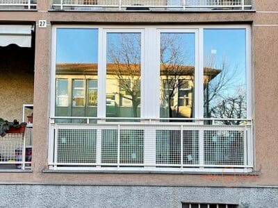 Veranda in PVC Brugmann con fissi inferiori e scorrevoli paralleli installata su balcone a Lodi. La struttura bianca con ampie vetrate riflette l'edificio circostante, creando uno spazio abitabile protetto e luminoso. Soluzione elegante per chiusura balconi con sistema scorrevole parallelo.