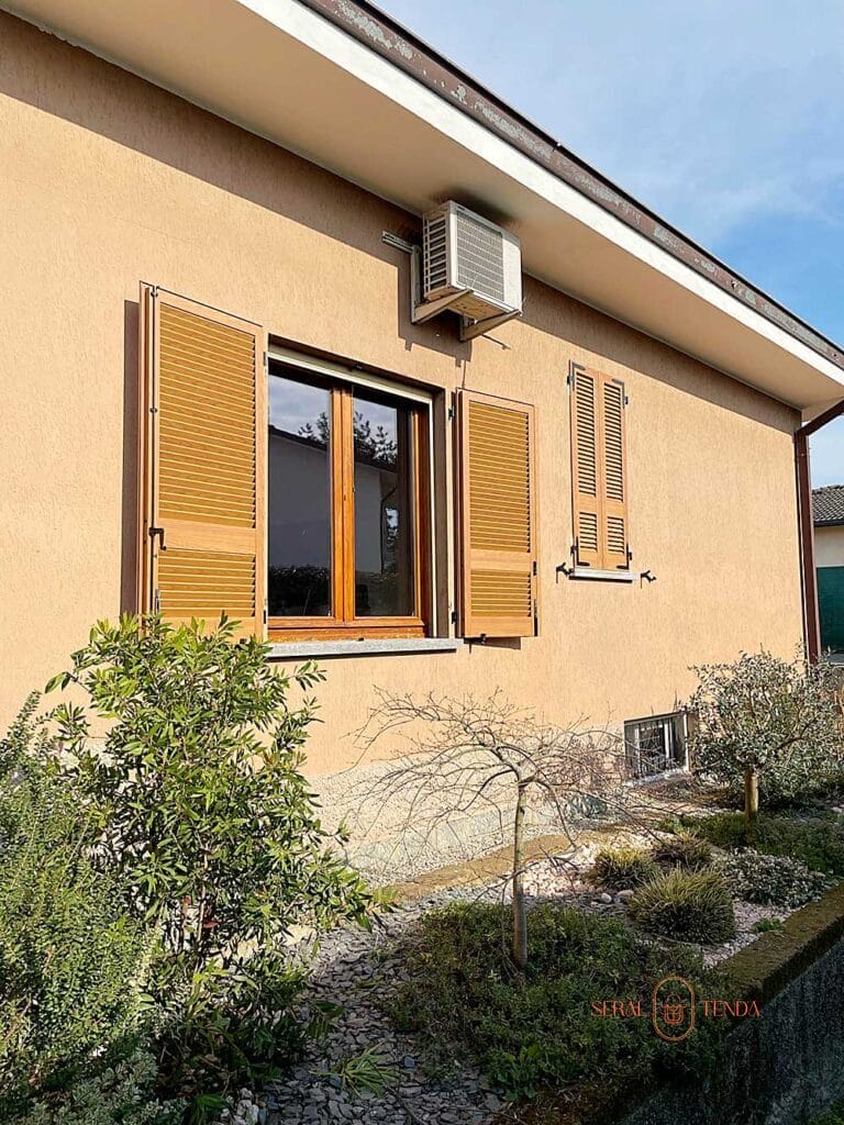 Esterno della casa color cuoio con persiane in legno Tinta Legno, un piccolo giardino e un condizionatore montato sopra la finestra: perfetto per un'accogliente Villa Montanaso Lombardo.