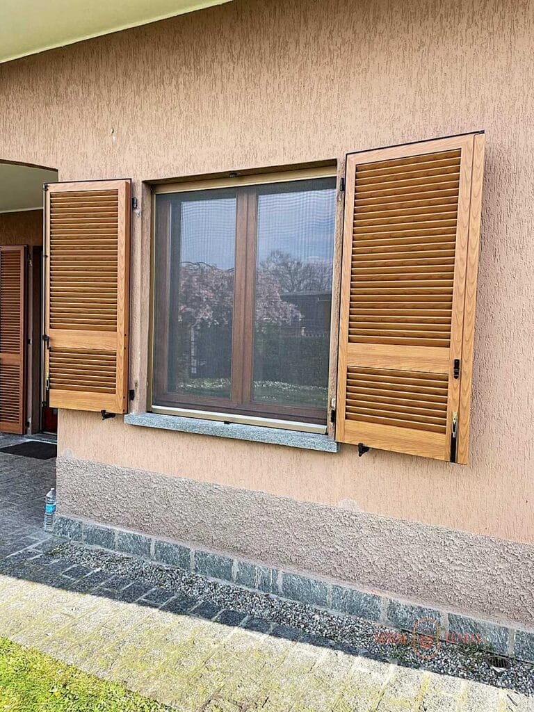 Una finestra con persiane aperte a lamelle fisse in tinta legno su una parete in stucco beige, sopra una bordura in pietra ed erba a Villa Montanaro Lombardo.