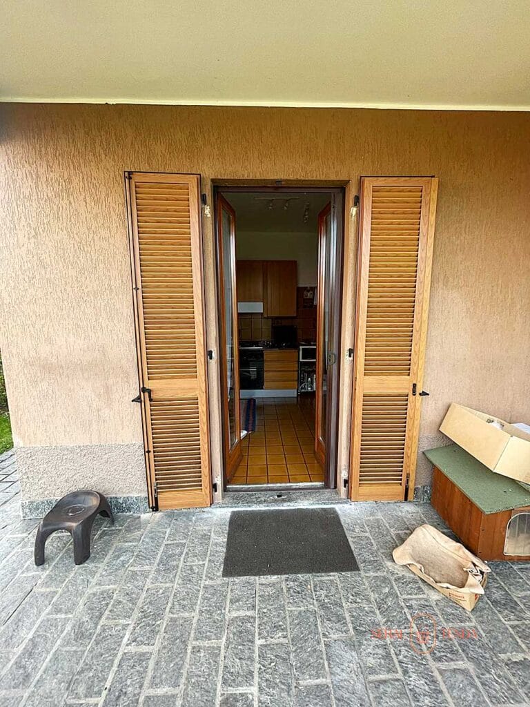 Una porta di legno aperta con persiane a lamelle fisse e tinta legno conduce alla cucina; scatole e sgabelli si trovano sul patio in pietra all'esterno di Villa Montanaso Lombardo.