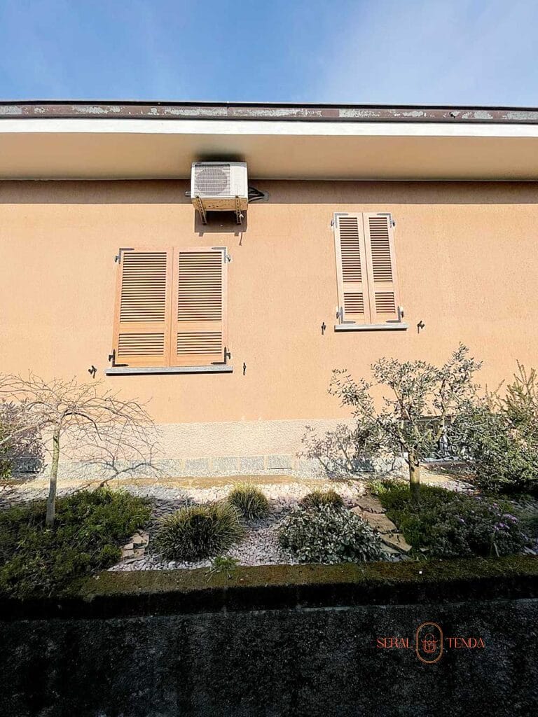 Parete color pesca a Villa Montanaso Lombardo, con due finestre Persiane a Lamelle Fisse, aria condizionata e un piccolo giardino con arbusti di fronte.