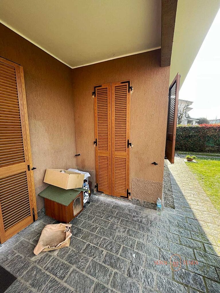 Un patio coperto con porte in legno rifinite in tinta legno, una scatola di cartone, una cuccia, un sacco di iuta e un giardino sullo sfondo a Villa Montanaso Lombardo.