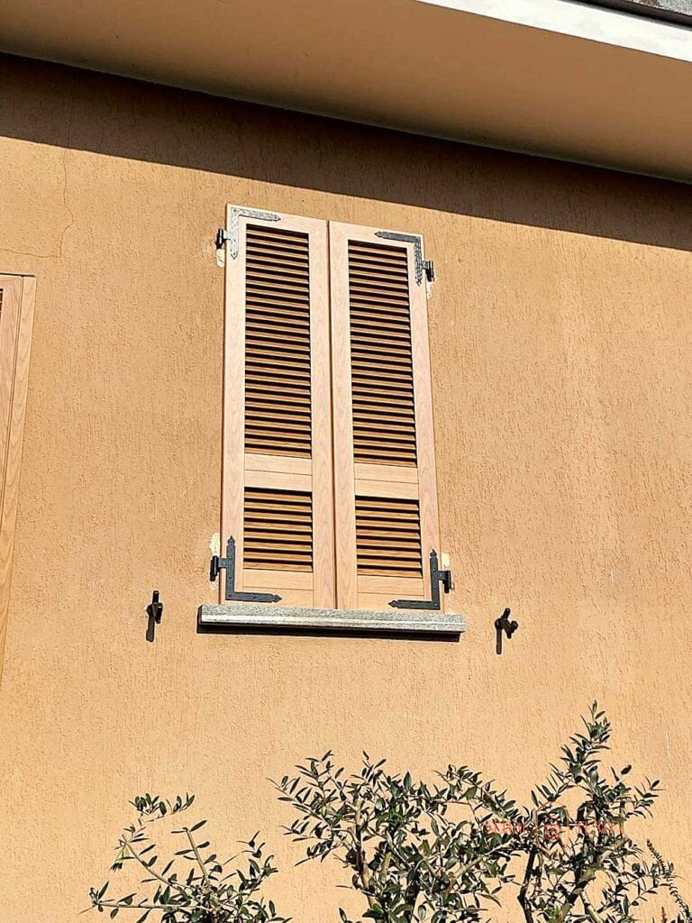 Persiane marrone chiaro a lamelle fisse su una parete beige, con la luce del sole che proietta ombre e una pianta in primo piano, evocano il caldo fascino di Villa Montanaro Lombardo.