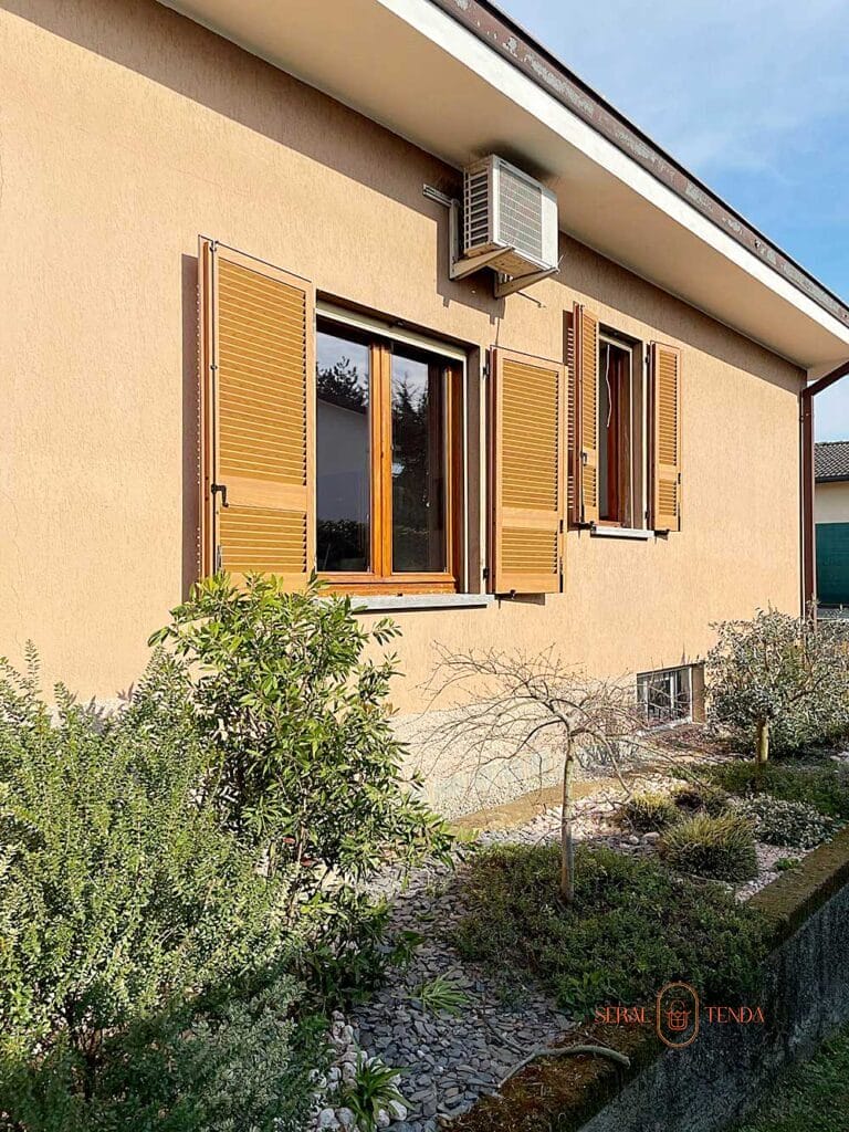 Una casa beige a Villa Montanaso Lombardo con persiane in legno Tinta Legno, aria condizionata e giardino con arbusti davanti.