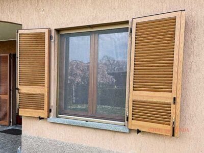 Una finestra con persiane a lamelle fisse e persiane in legno aperte su entrambi i lati, incastonata nella parete esterna beige, evoca il fascino classico di Villa Montanaro Lombardo.